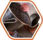 A person pouring a container of dark berries into a metal machine, illustrating the distilling and brewing of natural ingredients in the production of Raw Material Supplies.